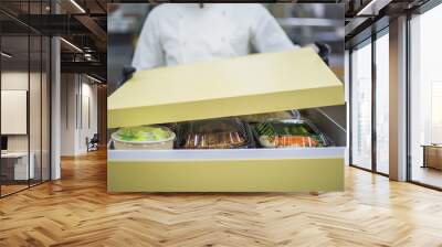 
A chef in black gloves and a white uniform is packing food delivery for customers in a large brown cardboard box. Wall mural