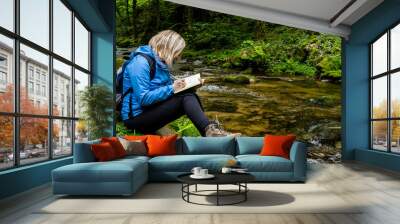 Young blond woman in a blue parka writing in her diary while sitting on a mossy rock by a fresh forest creek in mountains. Travel alone. Wall mural