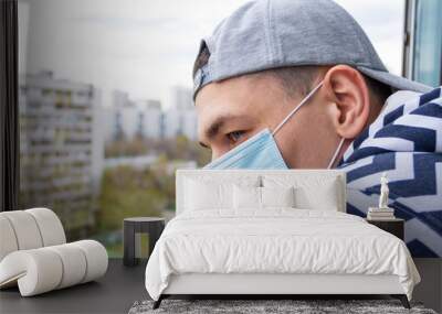 Young caucasian man in mask looking out to home balcony terrace who s quarantined in his apartment due to coronavirus covid19 pandemic. Wall mural