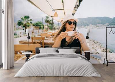 Young woman female in sunglasses and hat drinking coffee in a street outdoors seaside cafe restaurant coffee shop with scenery sea and mountains in the background. Hello summer holiday vacation. Wall mural