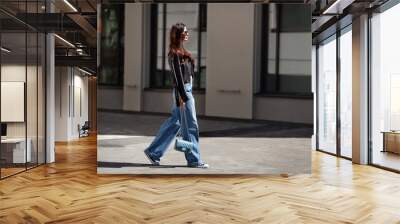 Full length beautiful young brunette woman Wearing black leather jacket and wide blue jeans, sneakers, sunglasses and small handbag with chain, walking street on sunny day Wall mural