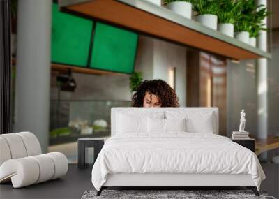 Woman with curly hair seats at vegetarian cafe or restaurant with green smoothie and plant meal on table, looking smartphone. Venues VEGAN sign displayed in background, healthy dining environment Wall mural