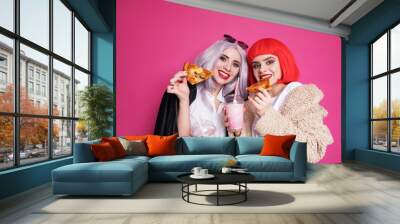 Two cute girl friends woman looking like teenagers standing on a pink background. They wearing colorful wigs and stylish casual outfits and eating pizza with milkshakes. Happy emotional portraits Wall mural