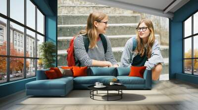 Outdoor portrait of two teenage girl sitting on stairs, wearing backbacks, talking to each other Wall mural