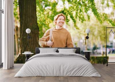 Outdoor sunny portrait of handsome happy young red-haired man in spring park, wearing brown fuzzy fleece sweater and backpack Wall mural