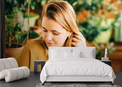 Romantic portrait of beautiful young woman wearing brown cotton dress, relaxing on the balcony between many green plants, holding cup of tea or coffee Wall mural