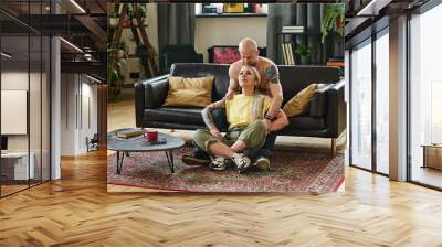 Attractive mature woman with long gray hair sitting on floor in living room leaning against her handsome husband Wall mural