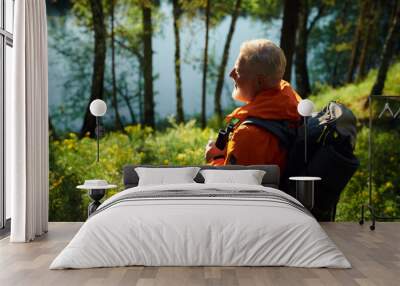 High angle view of senior male tourist wearing orange jacket with gray backpack looking at forest lake, copy space Wall mural