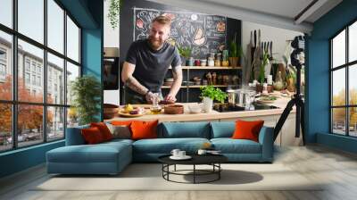 Joyful young adult Caucasian man with beard on face smiling at something while cooking dinner and filming process for blog Wall mural