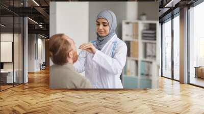 Nurse in hijab and in white coat examining the throat of patient during medical exam at hospital Wall mural