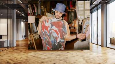 Person examining vibrant tie-dye shirt in cozy clothing store with a large window in background showcasing natural light. Shelves of various garments and unique decor surround them Wall mural