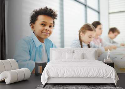 Portrait of cheerful African American boy sitting at school desk with his classmates looking at camera smiling, copy space Wall mural