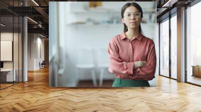 Portrait of confident young businesswoman in eyeglasses standing with arms crossed and looking at camera at office Wall mural