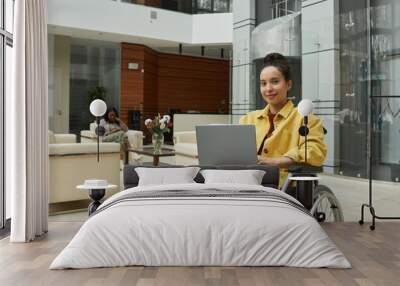 Portrait of young businesswoman sitting on wheelchair and smiling at camera she working online on laptop at office Wall mural