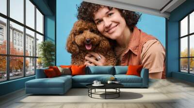 Portrait of young happy woman embracing her dog and smiling at camera isolated on blue background Wall mural