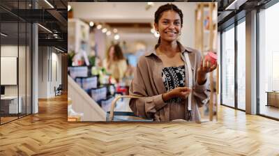 Portrait of young woman smiling at camera while choosing cream for herself in the store Wall mural