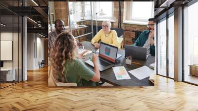 Rear view of young woman sitting at the table with computer and telling her colleagues about online work during a meeting at office Wall mural