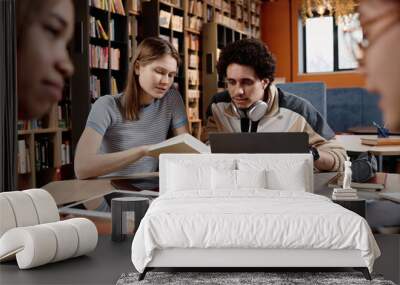 Selective focus shot of young male and female college students sitting in library doing school project using book and laptop Wall mural