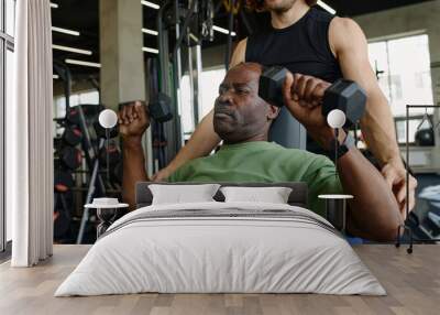 Senior black man lifting dumbbells at gym with unknown caucasian instructor standing behind helping Wall mural