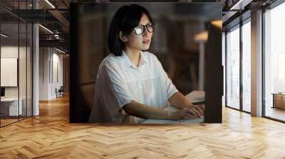 Serious Asian businesswoman wearing eyeglasses looking at computer monitor and typing she working till late Wall mural