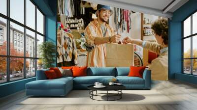 Smiling man wearing glasses and a hat handing shopping bag in a thrift shop with various clothes hanging in background Wall mural