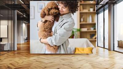 Vertical image of young woman holding dog in her arms and embracing it while standing in the room Wall mural