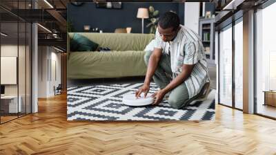 Young African American man sitting on floor in living room turning on robotic vacuum cleaner pressing button on it Wall mural