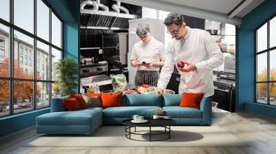 young chef in uniform choosing fresh vegetables for dish at table with his assistant making notes on Wall mural