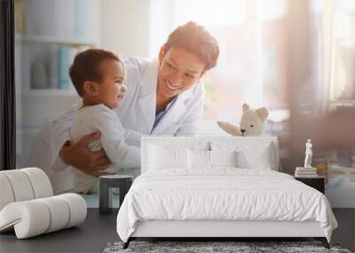 Young female doctor in white coat showing toy to her little patient and playing with him at hospital Wall mural