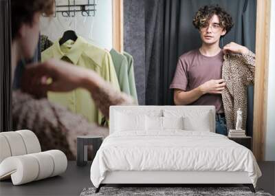 Young man standing in the fitting room in front of the mirror and trying on new shirt Wall mural