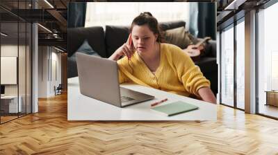 young woman with down syndrome sitting relaxed at table at home with laptop on it talking on phone w Wall mural