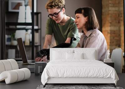 Young woman with prosthetic arm pointing at laptop and talking to her colleague during work at office Wall mural