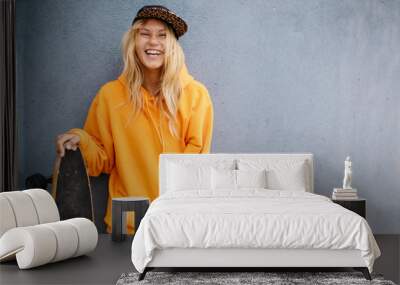 City portrait of positive young female wearing orange hoody and baseball cap holding skateboard. Grey blue wall on background Wall mural