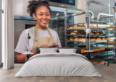 Close up of joyful African American young woman worker in apron stands in bakery shop, speking on smartphone and using tablet device. Small business concept. Wall mural