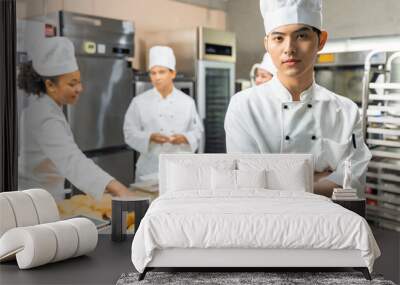 Smiling bakers looking at camera..Chef  baker in a chef dress and hat, cooking together in kitchen.Young asian man takes fresh baked cookies out of modern electric oven in kitchen. Wall mural