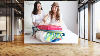 Young Asian woman packing clothes to the suitcase sitting on the bed. Prepairing for the summer vacations.Two women are planning a trip and helping to prepare luggage to travel abroad on a bed. Wall mural