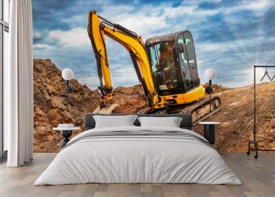 Mini excavator at the construction site on the edge of a pit against a cloudy blue sky. Compact construction equipment for earthworks. An indispensable assistant for earthworks. Wall mural