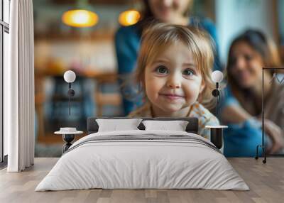 A close-up of a smiling toddler with big eyes and blonde hair, wearing a polka dot shirt. The background shows two adults sitting in a cozy, well-lit living room. Wall mural