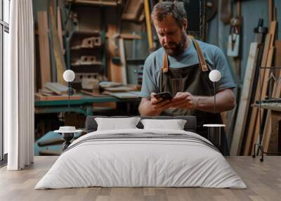 A craftsman in a woodworking workshop using a smartphone, surrounded by tools and wooden materials. Wall mural