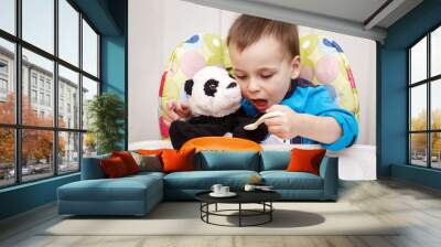 Close-up portrait of cute adorable little boy toddler two years old sitting in high chair in kitchen eating meal lunch dinner using fork and feeding his panda soft plush toy Wall mural