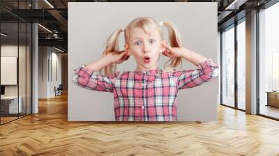 Closeup portrait of funny blonde Caucasian preschool girl making faces in front of camera. Child holding pulling dragging ears on light background. Kid expressing emotions. April fool day concept Wall mural