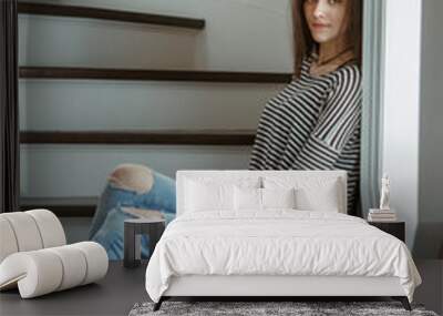 Closeup portrait of pensive Caucasian young beautiful woman model with messy long hair in ripped blue jeans and striped t-shirt sitting on stairs indoor looking in camera, toned with filters Wall mural