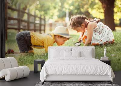 Cute adorable Caucasian girl and boy looking at plants grass in park through magnifying glass. Children friends siblings with loupe studying learning nature outside. Child education concept. Wall mural