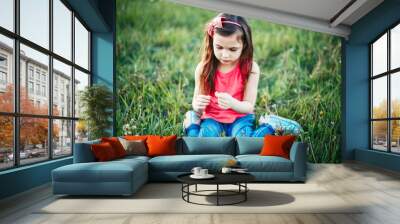 Cute adorable Caucasian girl picking flowers dandelions. Kid sitting in grass on meadow. Outdoors fun summer seasonal children activity. Child having fun. Happy childhood lifestyle. Wall mural