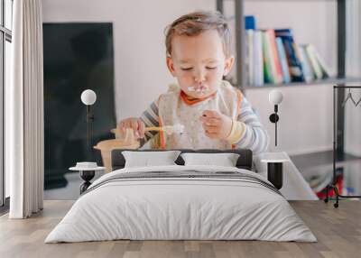 Cute Caucasian baby boy eating ripe berries and fruits with yogurt. Funny smiling child kid sitting in chair eating fresh berries and drinking water. Supplementary healthy food for toddler kid. Wall mural