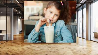 Funny smiling Caucasian preschool girl drinking milk shake with paper straw in cafe. Child kid eating lunch brunch in a restaurant patio enjoying food drink. Happy authentic childhood lifestyle. Wall mural