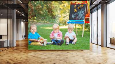 Group of three white Caucasian toddler children kids boys and girl sitting outside in summer autumn park by drawing easel with books, reading studying learning, back to school Wall mural