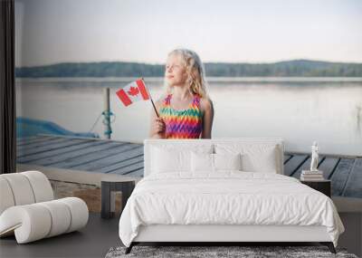 Happy Caucasian girl sitting on pier by lake and waving Canadian flag. Smiling child holding Canada flag by water. Kid celebrating Canada Day holiday on first day of July outdoor Wall mural