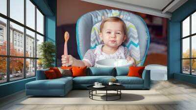 Portrait of cute adorable Caucasian child kid girl sitting in high chair eating cereal with spoon. Everyday lifestyle. Candid real authentic moment Wall mural