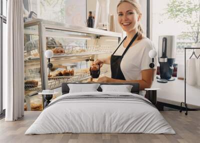 Portrait of white Caucasian beautiful barista woman taking muffin pastry from shop-window. Person at work place, small business concept Wall mural